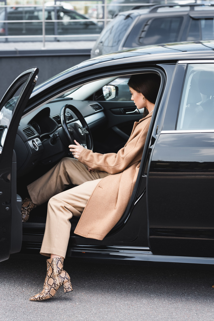 молодая женщина в стильном наряде сидит в машине с открытой дверью и держит руль - Фото, изображение