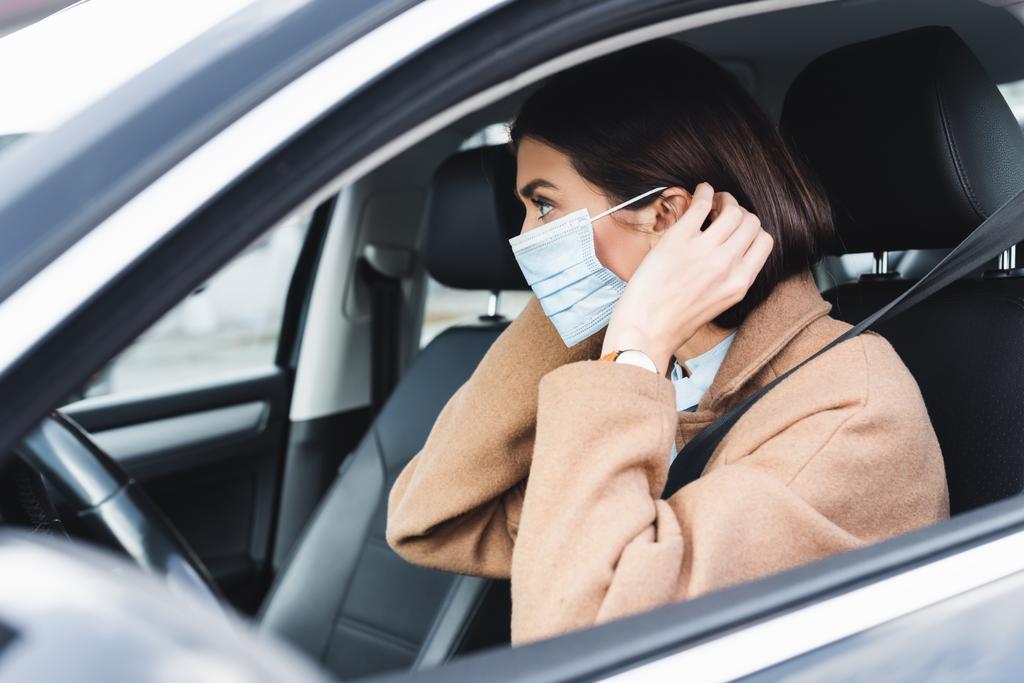 mujer joven en traje de otoño ponerse máscara médica mientras está sentado en el coche en primer plano borrosa - Foto, Imagen