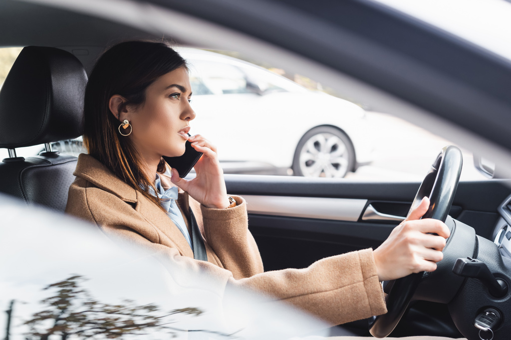 Trençkotlu şık bir kadının cep telefonuyla konuşurken ön planda bulanık bir araba sürüşünün yan görüntüsü - Fotoğraf, Görsel