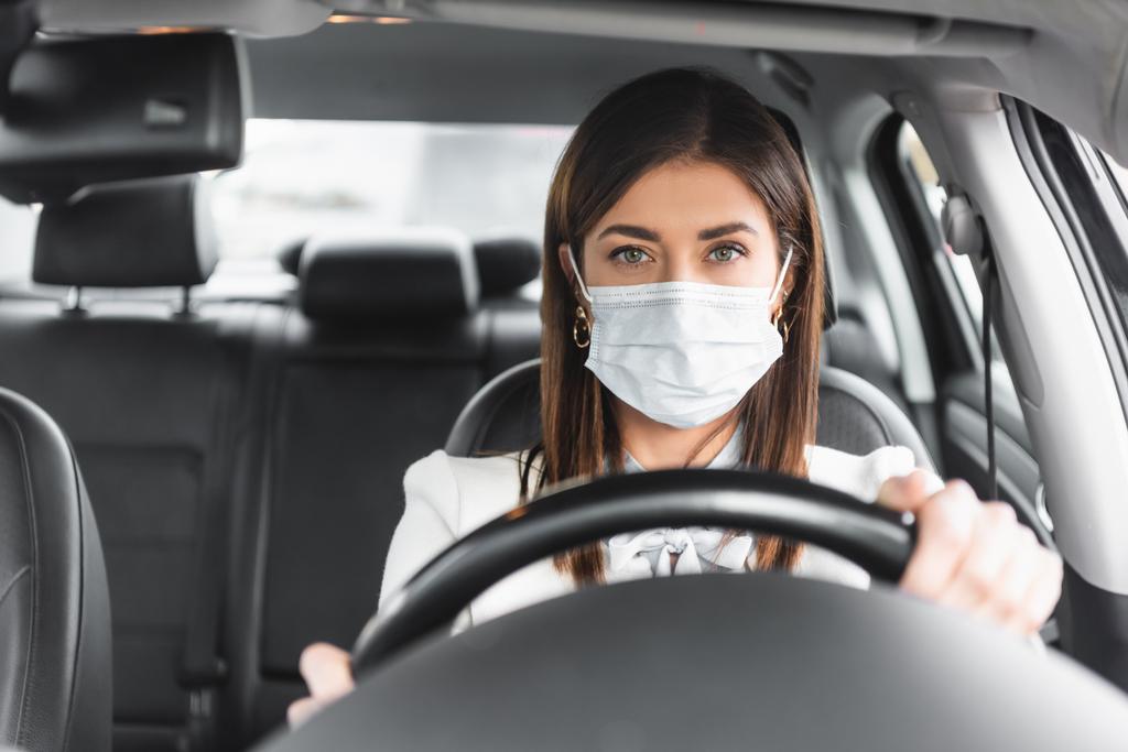 женщина в медицинской маске смотрит в камеру во время вождения автомобиля на размытом переднем плане - Фото, изображение