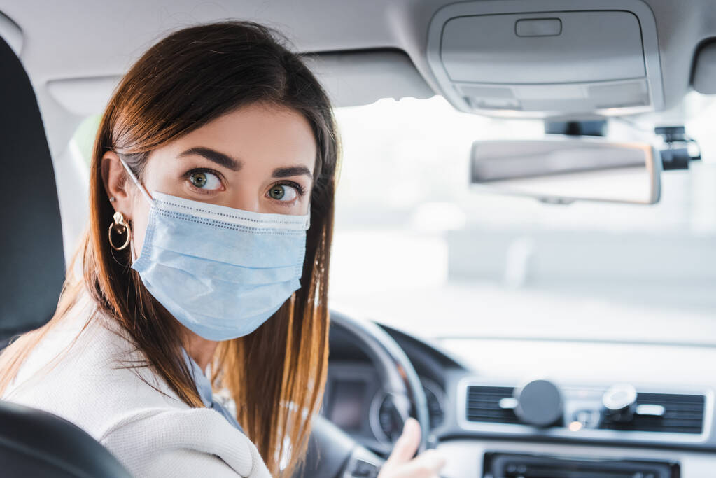Nuori nainen lääketieteellisessä naamiossa etsii taaksepäin ajaessaan autoa hämärtyneellä taustalla - Valokuva, kuva