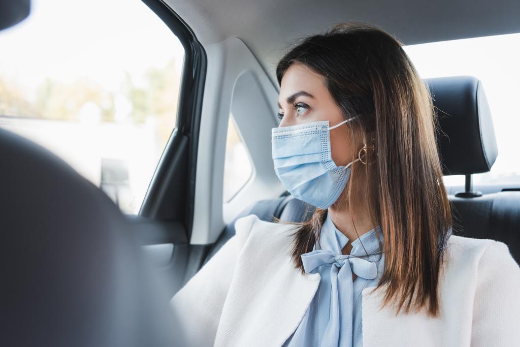 κομψή γυναίκα με ιατρική μάσκα κοιτάζοντας έξω από το παράθυρο, ενώ κάθεται στο πίσω κάθισμα του αυτοκινήτου σε θολή φόντο - Φωτογραφία, εικόνα
