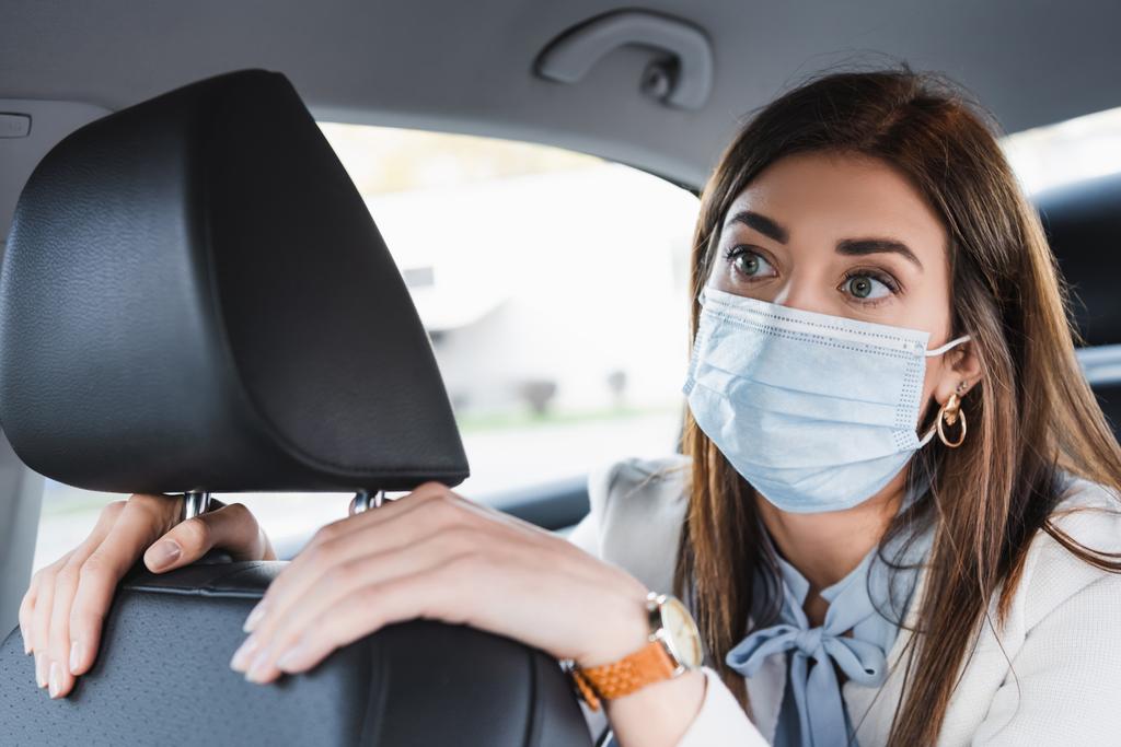 mujer joven con máscara médica tocando el asiento delantero y mirando hacia el futuro mientras monta en coche - Foto, imagen