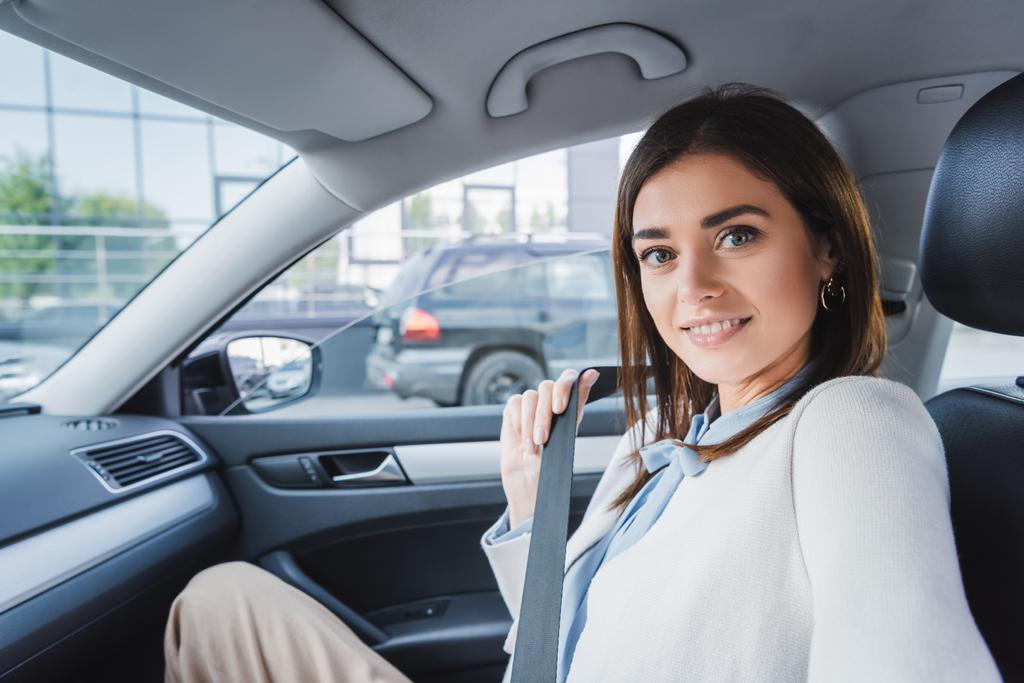 elegant woman smiling at camera while sitting in car and fixing seatbelt - Photo, Image