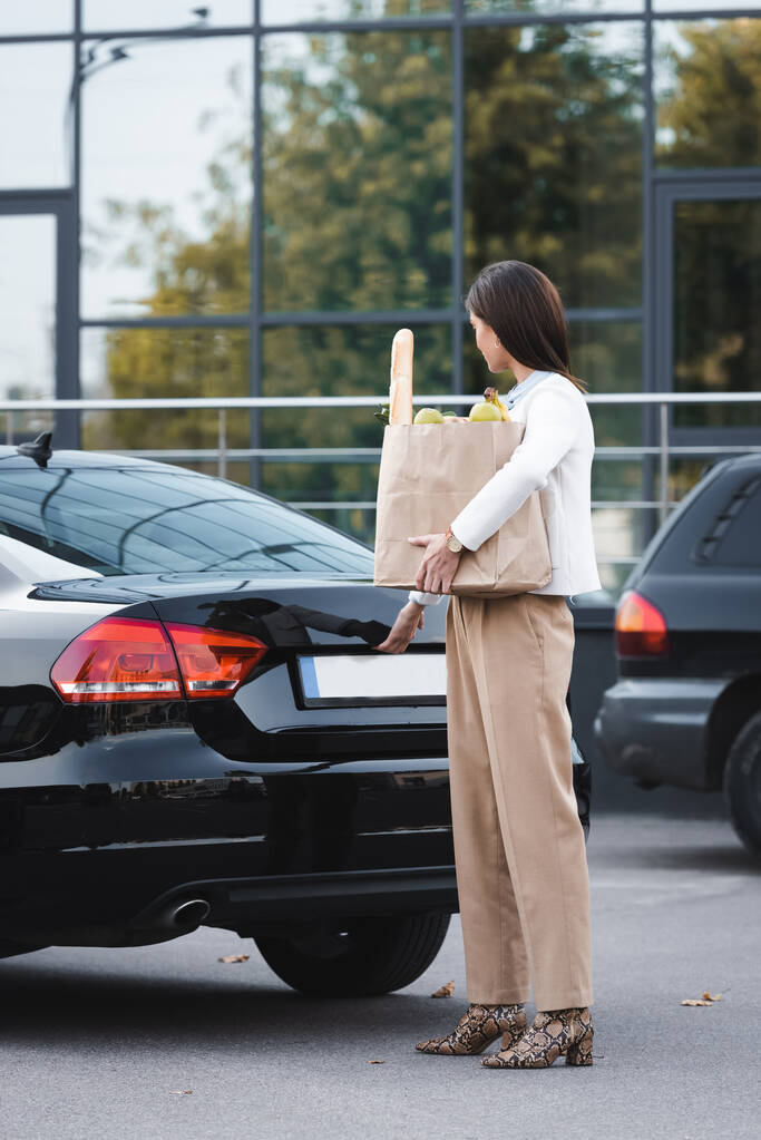 Frau öffnet Kofferraum von schwarzem Auto und hält Einkaufstasche mit Lebensmitteln in der Hand - Foto, Bild