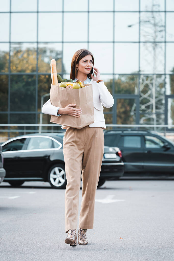 stijlvolle vrouw praten op smartphone tijdens het wandelen langs parkeerplaats met voedsel in boodschappentas - Foto, afbeelding
