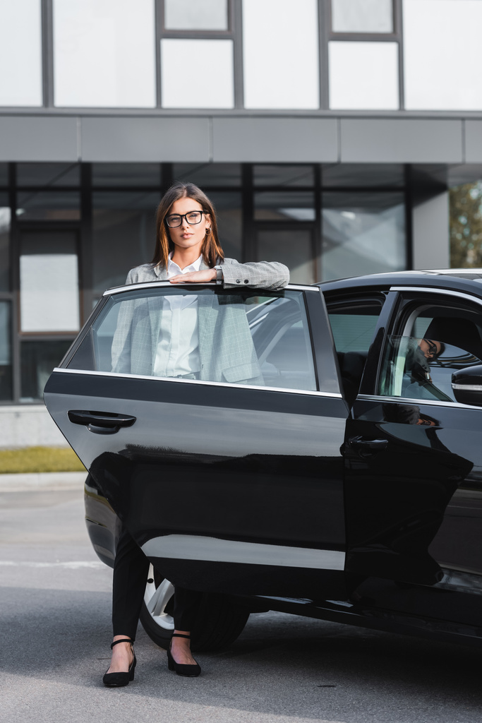 confiante, mulher de negócios elegante em óculos de pé perto de carro preto com porta aberta - Foto, Imagem