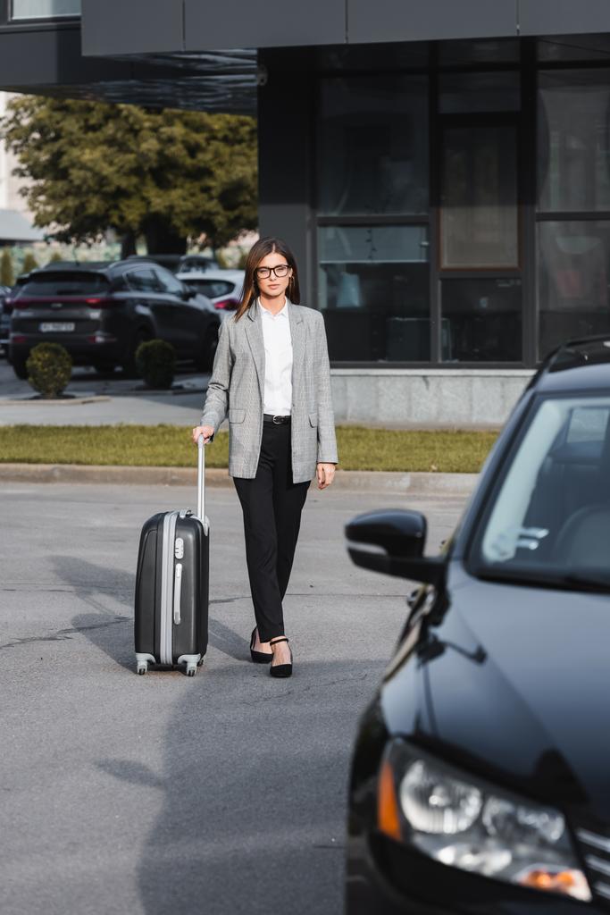 κομψή επιχειρηματίας περπάτημα με βαλίτσα κοντά στο μαύρο αυτοκίνητο σε θολή πρώτο πλάνο - Φωτογραφία, εικόνα