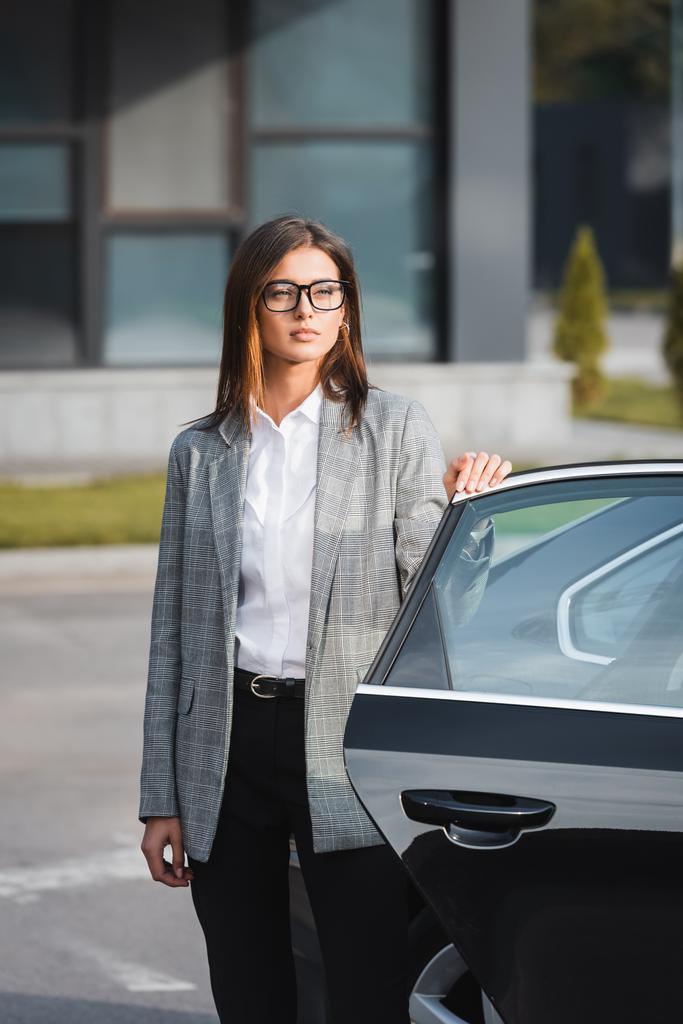 jonge zakenvrouw in formele kleding en bril staan in de buurt van open auto - Foto, afbeelding