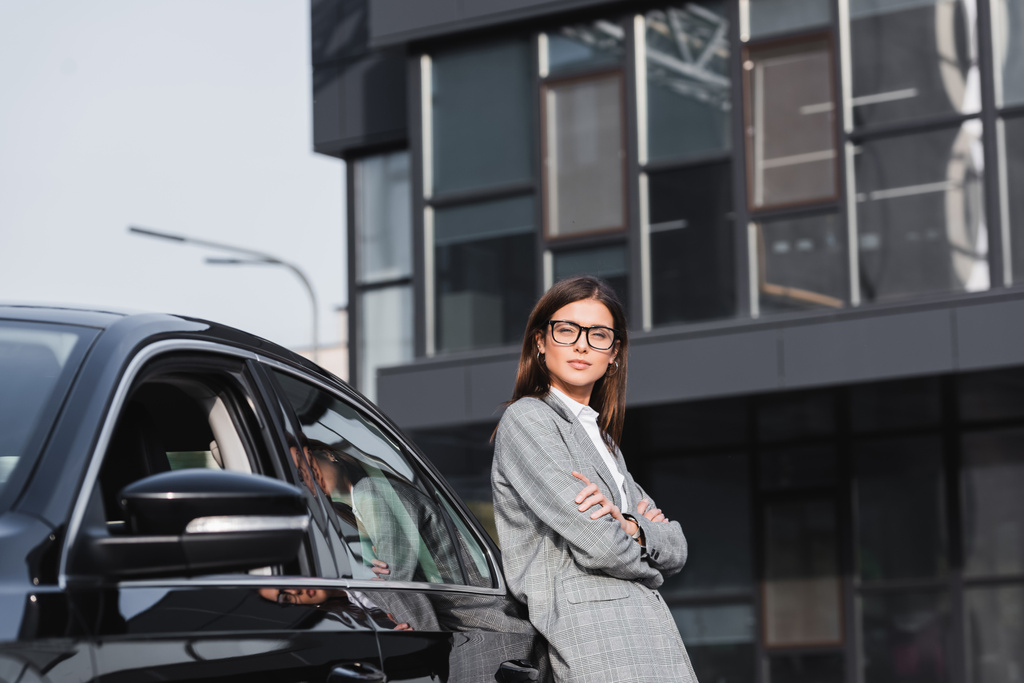 молодая стильная деловая женщина, опирающаяся на черный автомобиль, стоя со скрещенными руками - Фото, изображение