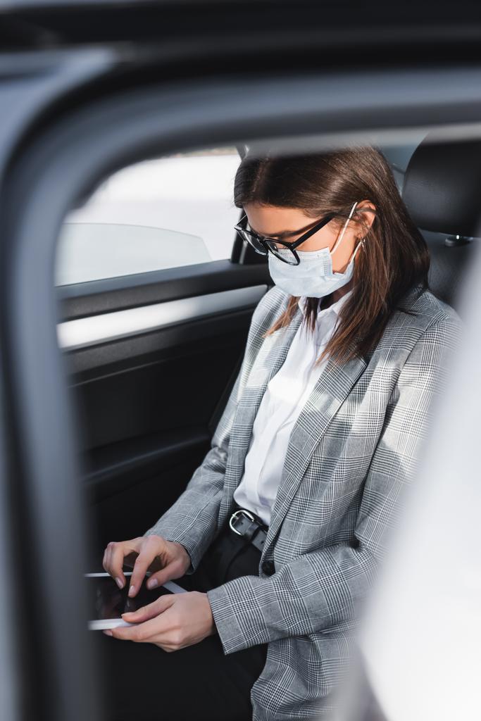 mujer de negocios en máscara médica utilizando tableta digital mientras se monta en coche en primer plano borrosa - Foto, imagen