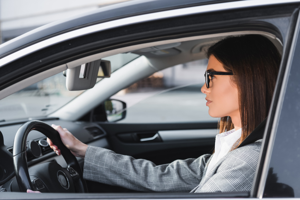 vista lateral de la mujer de negocios en gafas de vista mirando hacia el futuro mientras conduce el coche - Foto, Imagen