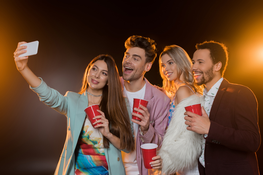 gruppo di amici multiculturali felici prendendo selfie mentre tenendo bicchieri di plastica sul nero - Foto, immagini
