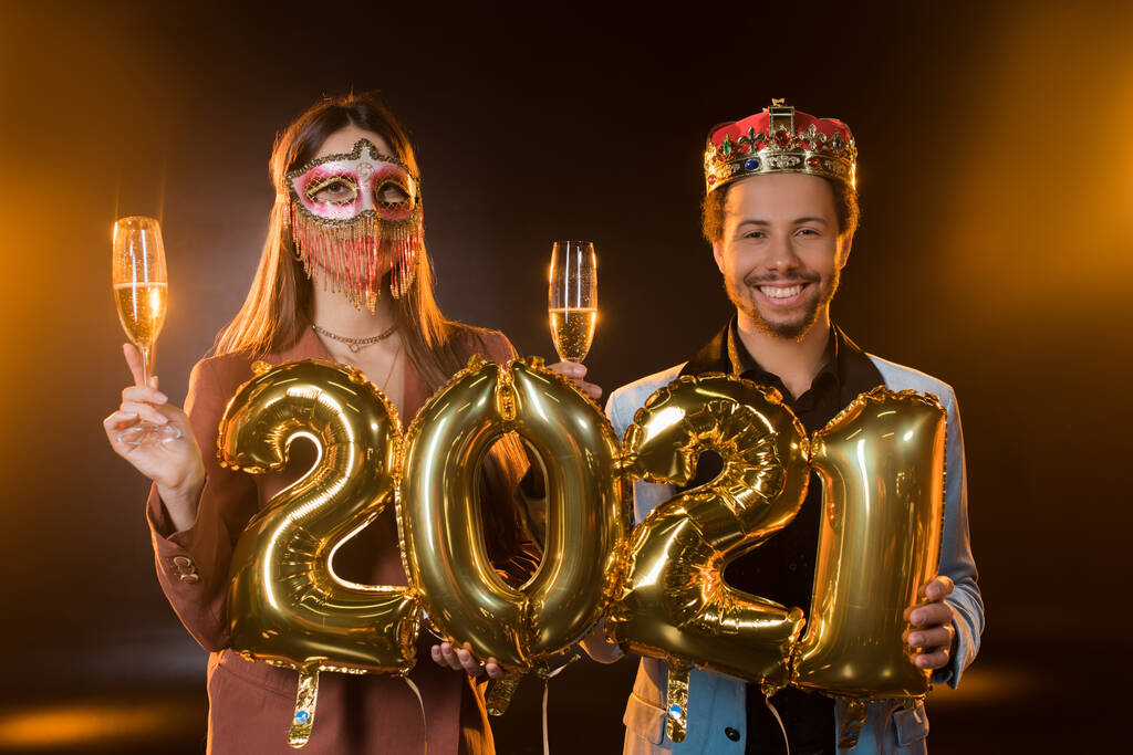 homem americano africano feliz na coroa segurando balões com 2021 números perto de mulher em máscara de carnaval com copo de champanhe em preto  - Foto, Imagem