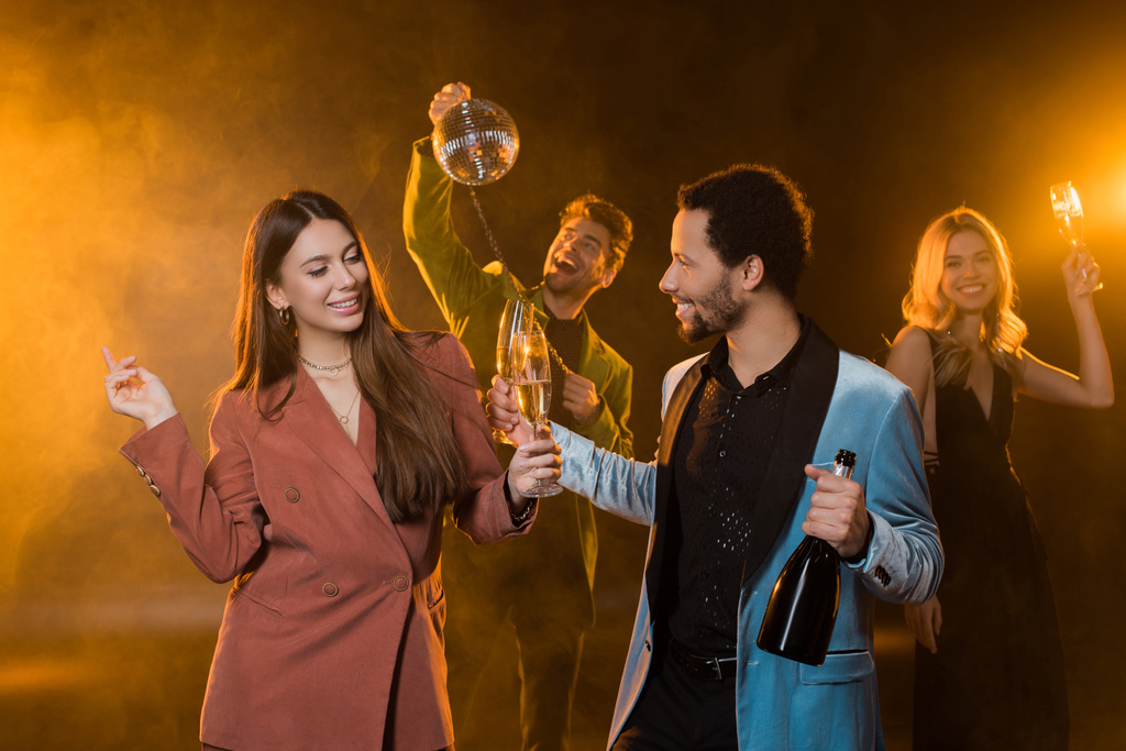 szczęśliwa wielokulturowa para trzymająca kieliszki szampana i butelki w pobliżu wesołych przyjaciół na rozmazanym i czarnym tle - Zdjęcie, obraz