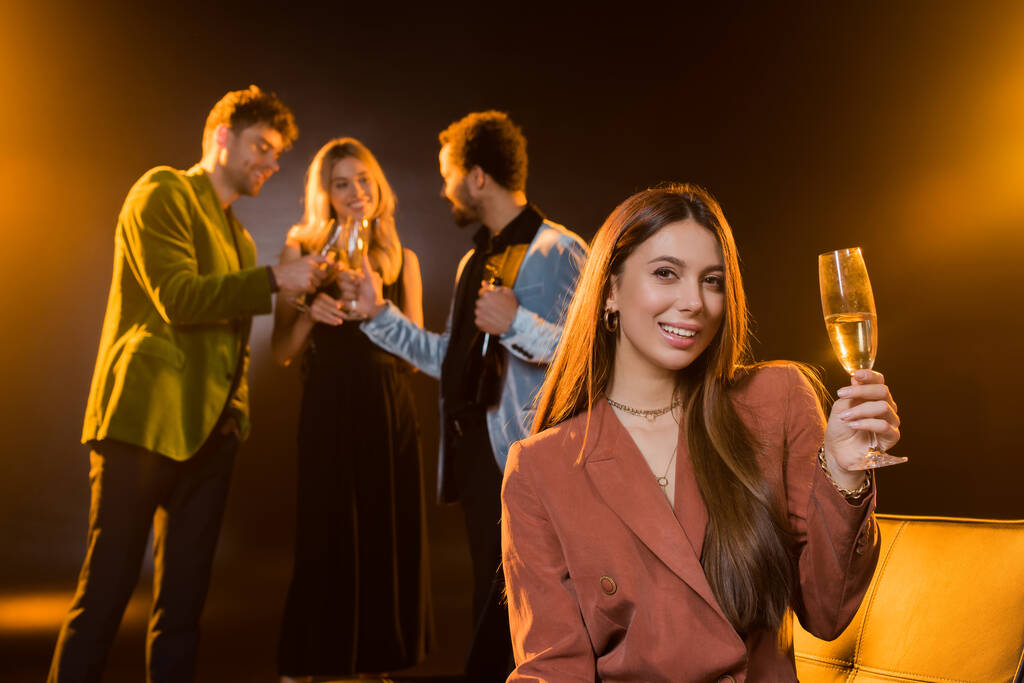 szczęśliwa brunetka kobieta siedzi na kanapie i trzyma szklankę szampana w pobliżu wielokulturowych przyjaciół na niewyraźne czarne tło - Zdjęcie, obraz