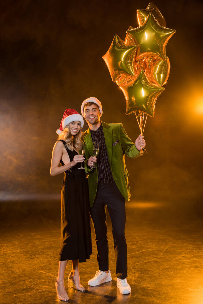 volledige lengte van het glimlachende paar in santa hoeden met glazen met champagne en ballonnen op zwart - Foto, afbeelding