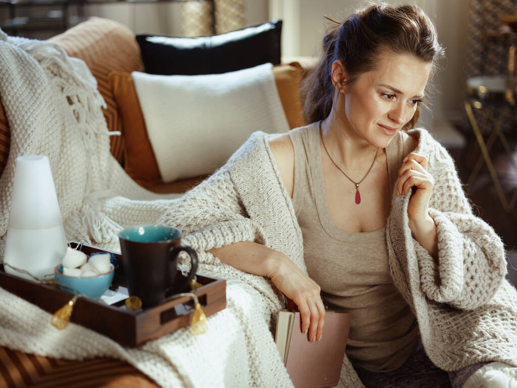 zamyšlená moderní žena v pleteném útulném svetru s knihou, podnosem, aromatickou lampou, hrnkem a pečivem v moderním obývacím pokoji za slunečného zimního dne. - Fotografie, Obrázek