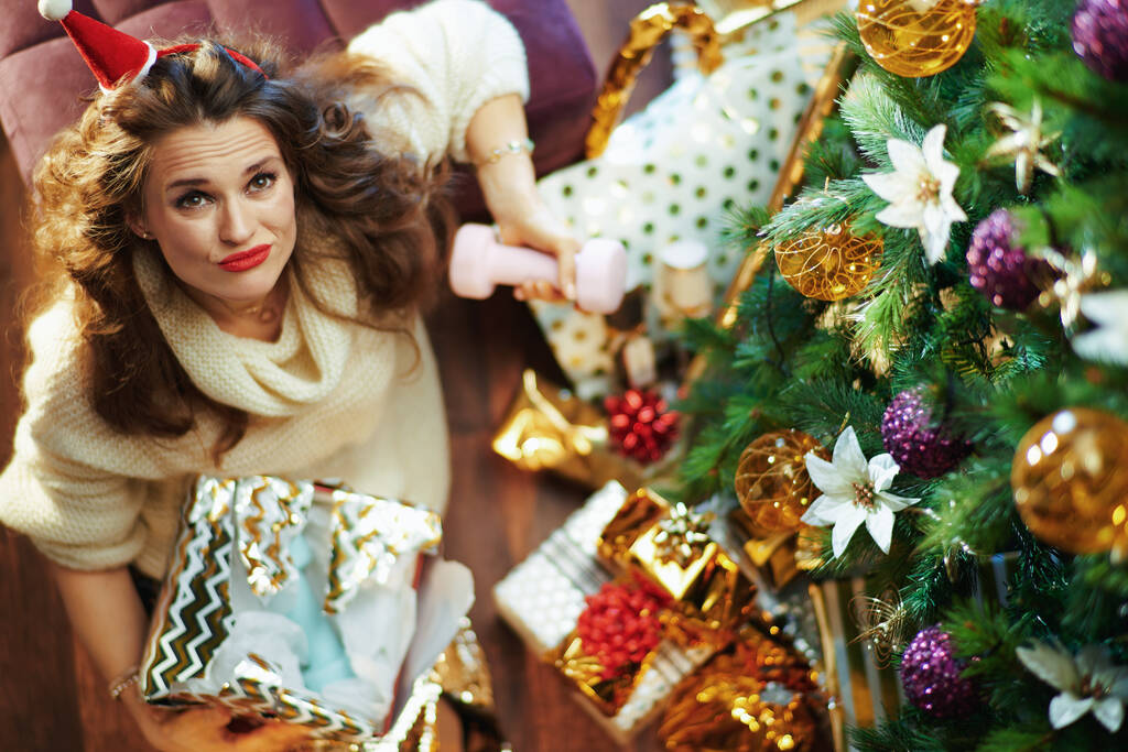 Merry Christmas. Upper view of unhappy trendy female in gold sequin skirt and sweater with dumbbells in christmas present box near Christmas tree and gift boxes. - Photo, Image