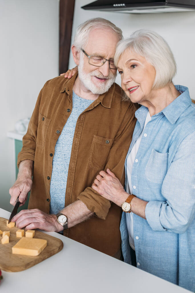 χαμογελαστή ηλικιωμένη σύζυγος αγκαλιάζει τον σύζυγο κόβοντας τυρί σε ξύλο κοπής στην κουζίνα  - Φωτογραφία, εικόνα
