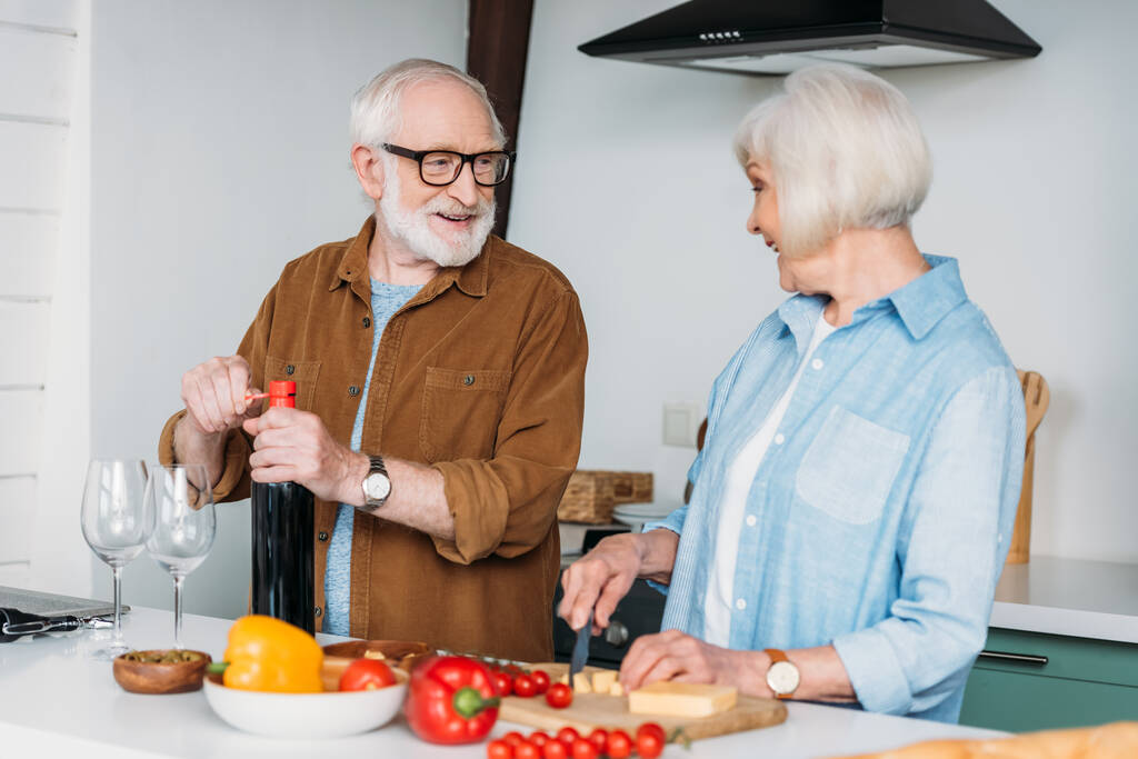 glücklicher älterer Mann schaut Frau beim Käseschneiden zu, während er eine Flasche Wein in der Küche öffnet - Foto, Bild