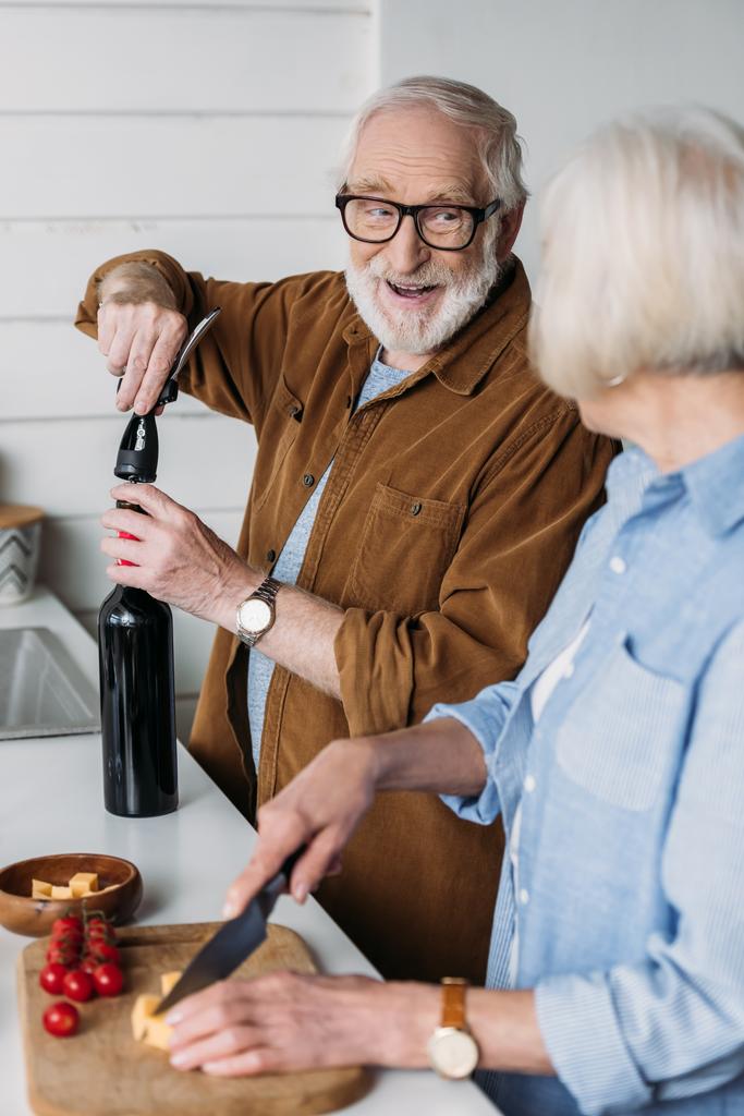 feliz marido anciano mirando a la esposa al abrir la botella de vino con sacacorchos en la cocina en primer plano borrosa - Foto, imagen