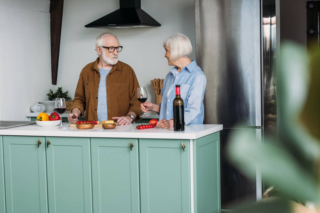 starszy mężczyzna patrząc na kobietę z kieliszkiem wina podczas gotowania obiadu w kuchni na zamazanym pierwszym planie - Zdjęcie, obraz