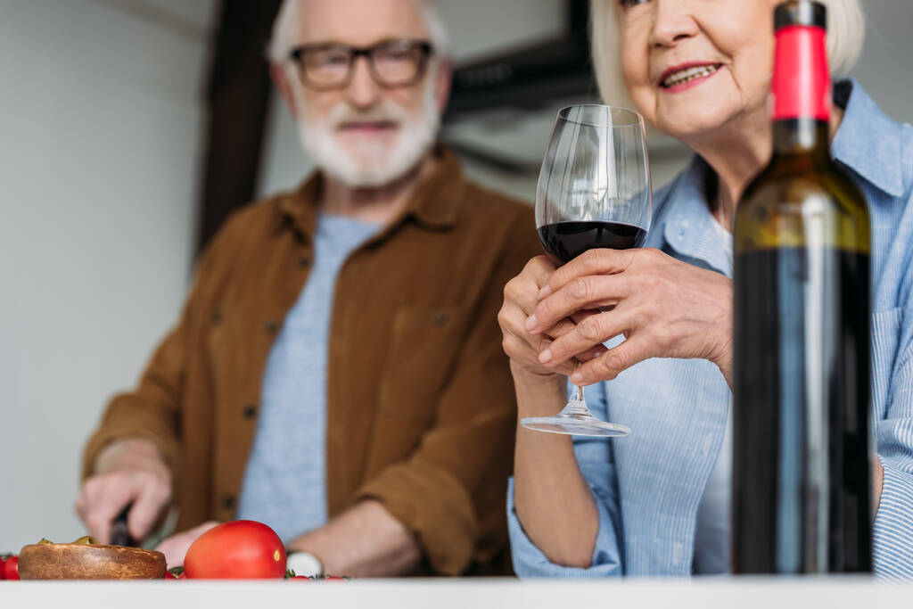 Seniorin hält Weinglas neben Flasche mit verschwommenem Mann im Hintergrund - Foto, Bild