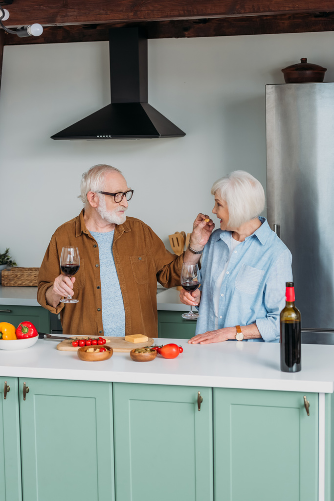 Lächelnder älterer Mann mit Weinglas füttert Frau mit Käsestück in Küche - Foto, Bild