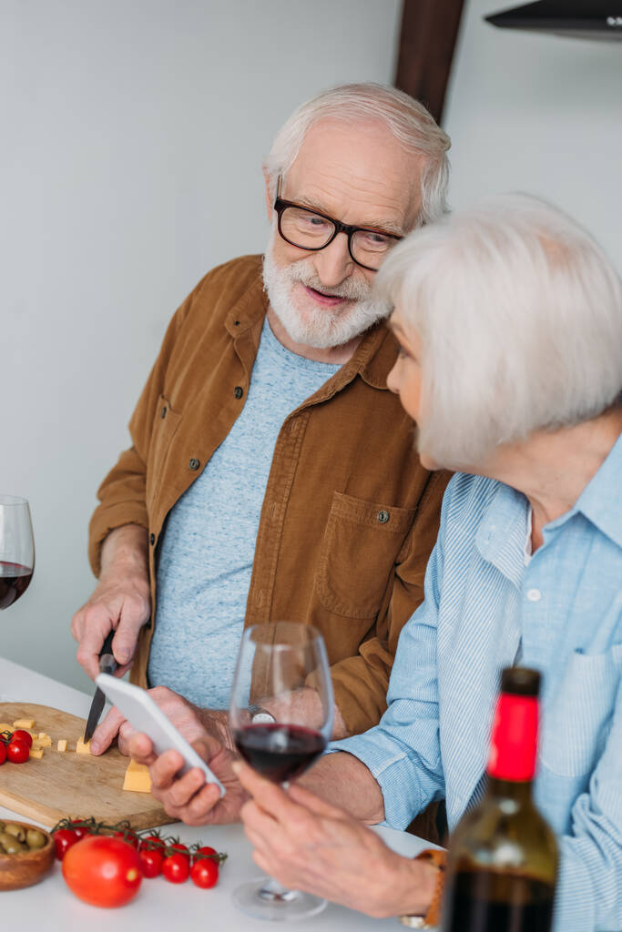 χαμογελαστός ηλικιωμένος σύζυγος που κοιτάζει τη σύζυγο ενώ κόβει τυρί σε ξύλο κοπής κοντά σε λαχανικά σε θολή πρόσθια επιφάνεια - Φωτογραφία, εικόνα