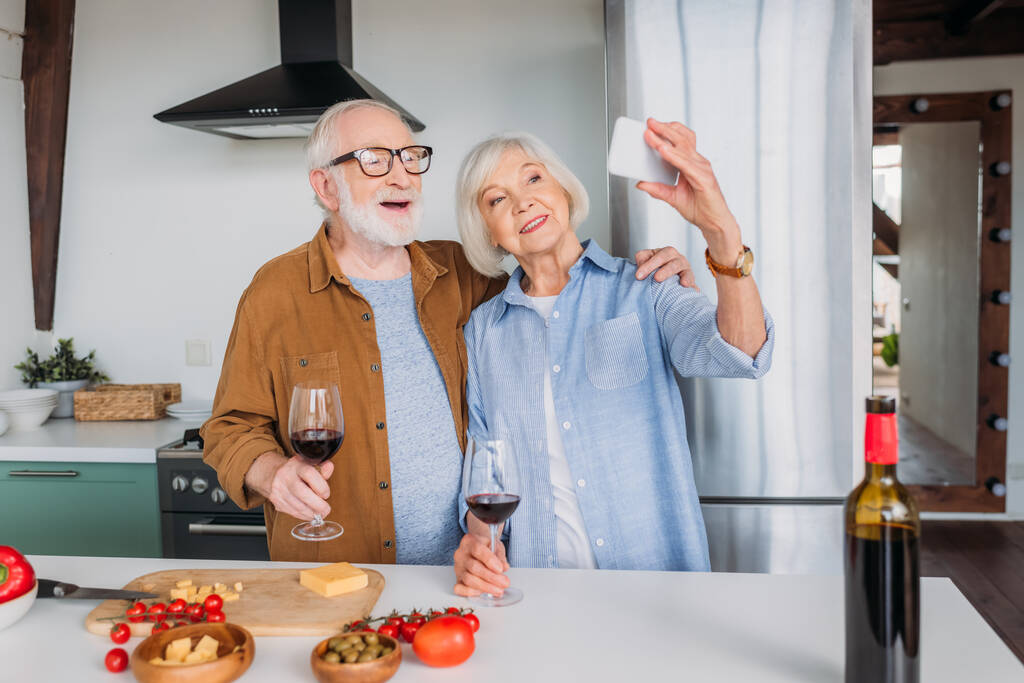 幸せな高齢者のカップルワイングラスはキッチンで食べ物とテーブルの近くで自撮り - 写真・画像