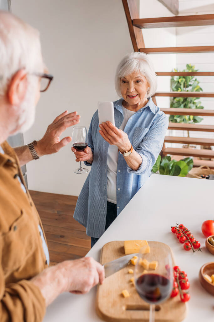 femme âgée souriante avec smartphone prenant la photo du mari en train de cuisiner le dîner au premier plan flou dans la cuisine - Photo, image