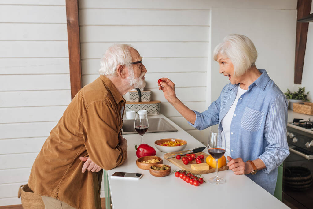 Seitenansicht der glücklichen Seniorin, die ihren Mann mit Kirschtomaten füttert, am Tisch mit Essen, Weingläsern und Smartphone in der Küche - Foto, Bild