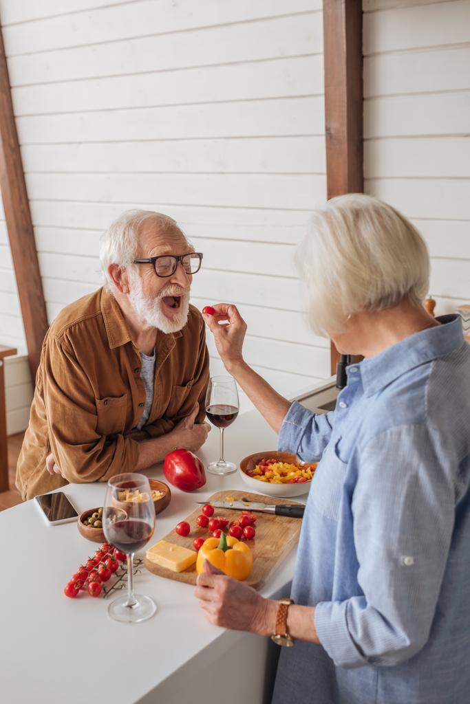 zpět pohled na starší ženy krmení šťastný manžel s cherry rajče v blízkosti stolu s jídlem v kuchyni - Fotografie, Obrázek