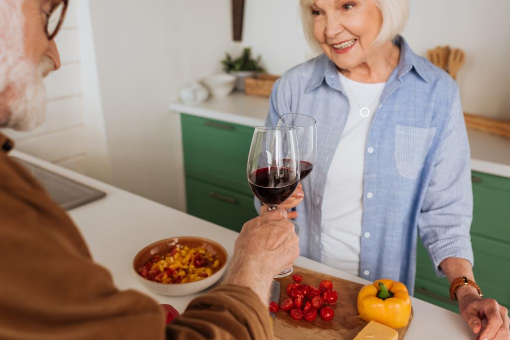 szczęśliwy starsza para toasting z kieliszkami wina w pobliżu stołu z jedzeniem na zamazanym pierwszym planie - Zdjęcie, obraz