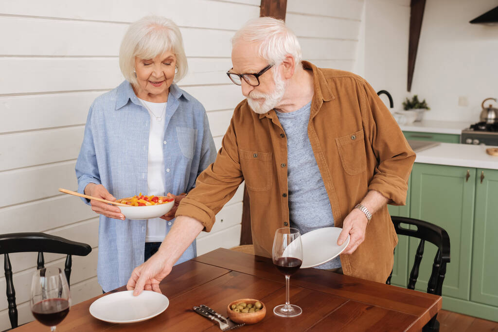 χαμογελαστό ηλικιωμένο ζευγάρι σερβίρει τραπέζι με πιάτα και σαλάτα στην κουζίνα σε θολή φόντο - Φωτογραφία, εικόνα