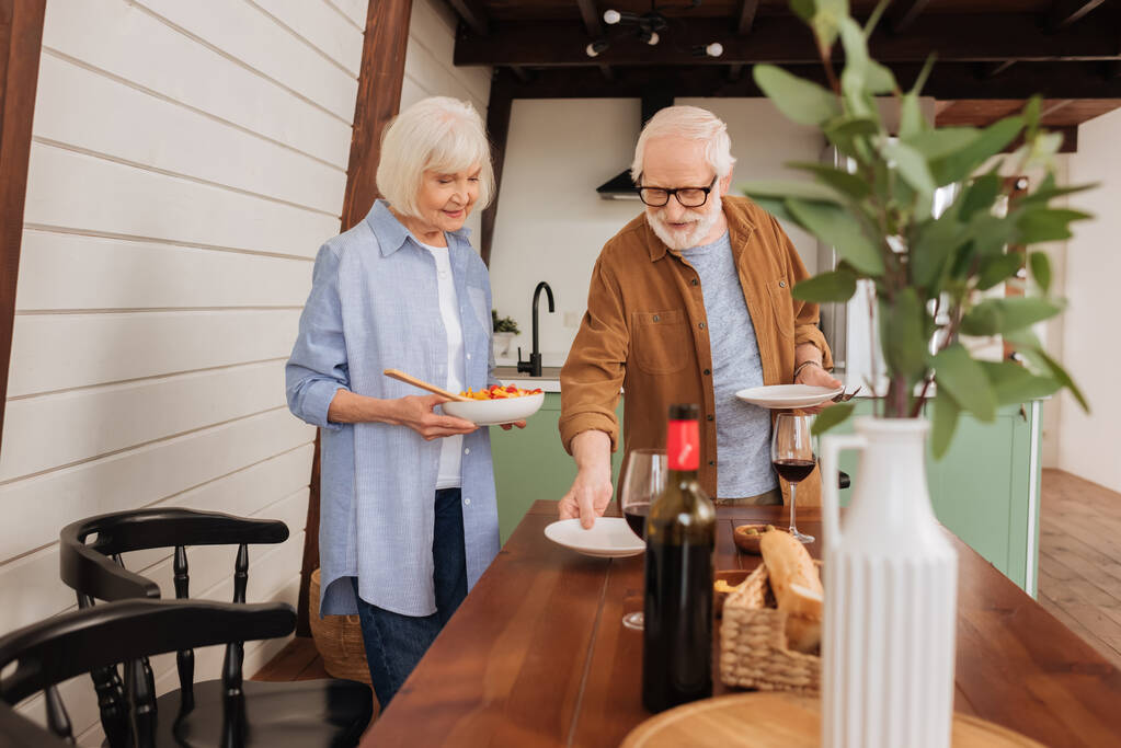 table de service de couple senior souriant avec assiettes et salade dans la cuisine avec plante décorative floue au premier plan - Photo, image