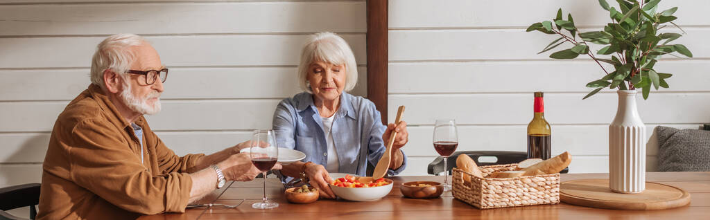 sorrindo esposa sênior com espátula servindo salada para marido à mesa com jantar vegetariano na cozinha, banner - Foto, Imagem