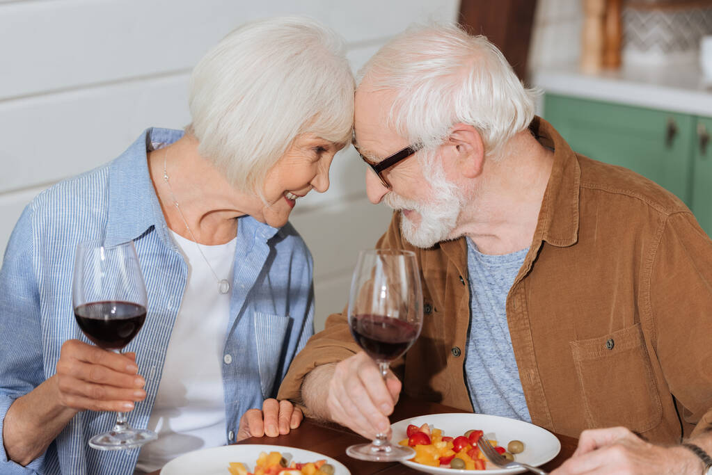 feliz pareja de ancianos con copas de vino mirándose mientras están sentados en la mesa con la cena vegetariana en un fondo borroso - Foto, Imagen