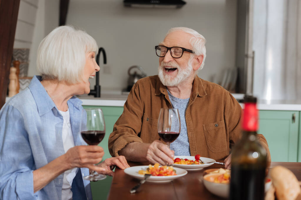 счастливая пожилая пара с бокалами вина смеются, глядя друг на друга за столом с ужином на размытом переднем плане - Фото, изображение