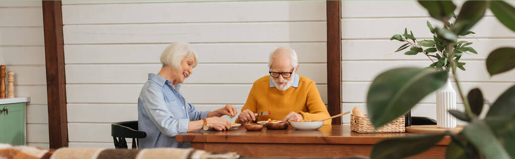 glückliches Seniorenpaar beim vegetarischen Abendessen, während es am Tisch sitzt, auf verschwommenem Vordergrund in der Küche, Banner - Foto, Bild