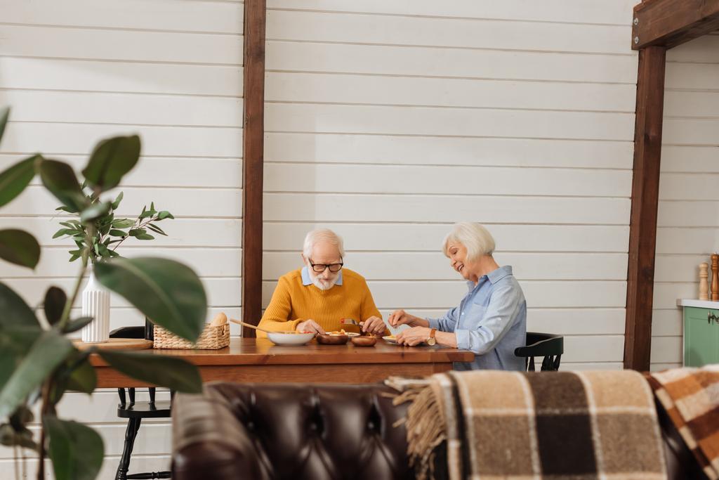 sonriente pareja de ancianos cenando en la mesa en primer plano borroso en la cocina - Foto, Imagen