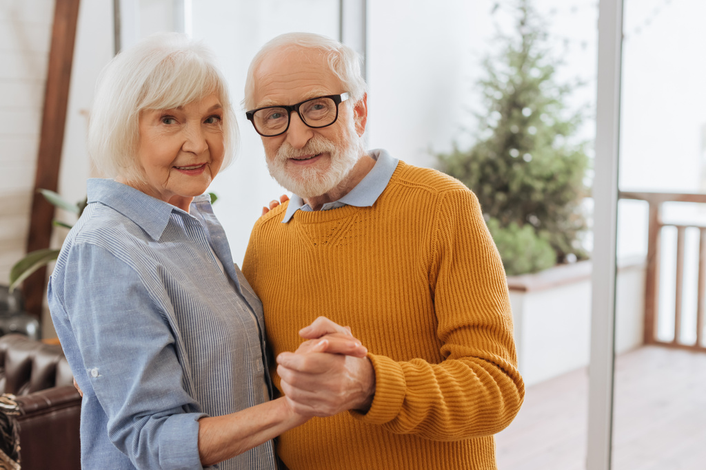 smiling elderly couple looking at camera while dancing together at home on blurred background - Photo, Image