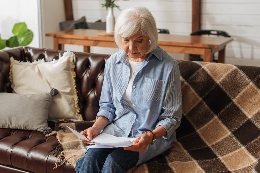 senior vrouw op zoek naar rekeningen terwijl zitten op de bank op wazig achtergrond in de woonkamer  - Foto, afbeelding