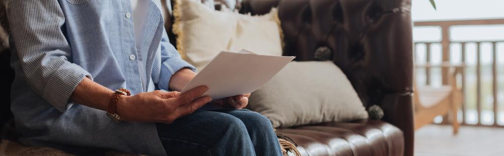 ostříhaný pohled na starší ženu držící účty, zatímco sedí na gauči na rozmazaném pozadí doma, prapor - Fotografie, Obrázek