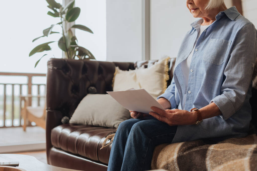 cropped view of elderly woman holding bills while sitting on couch on blurred background at home - Photo, Image