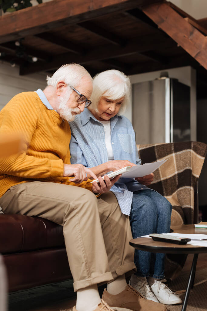 idős férj számológép közelében feleség nézi számlák kanapén homályos háttér a nappaliban - Fotó, kép