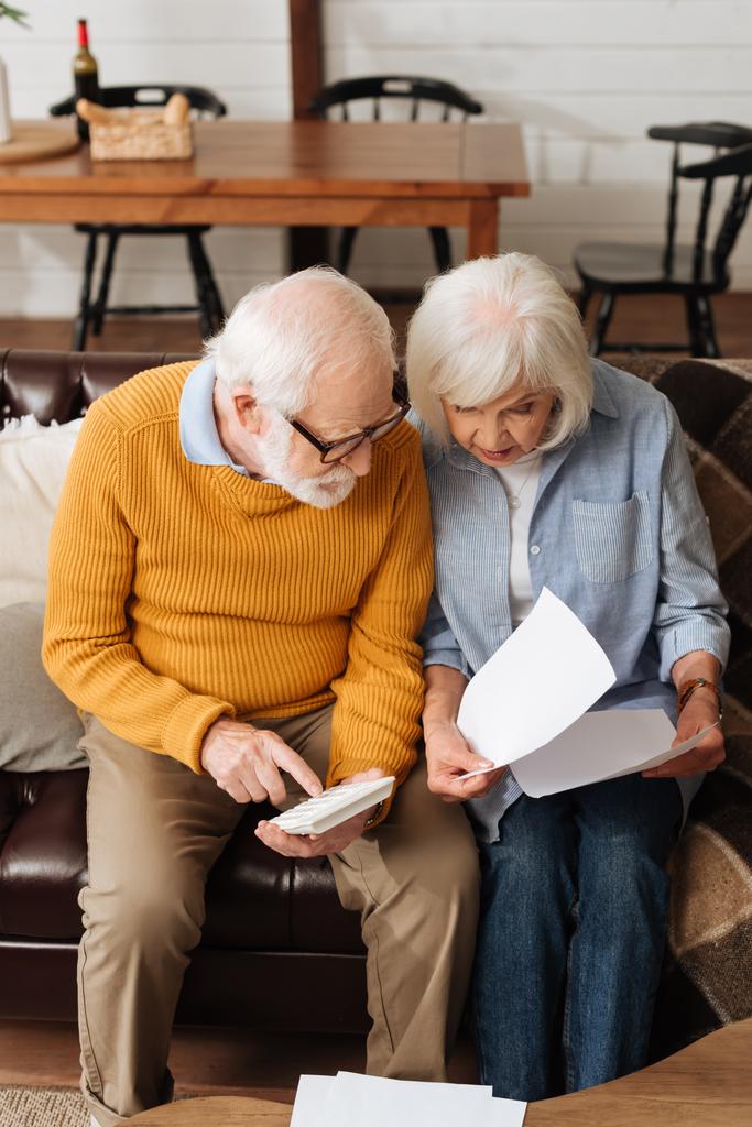 Senior Ehemann mit Taschenrechner schaut Rechnungen in der Nähe Frau auf Couch auf verschwommenem Hintergrund zu Hause - Foto, Bild