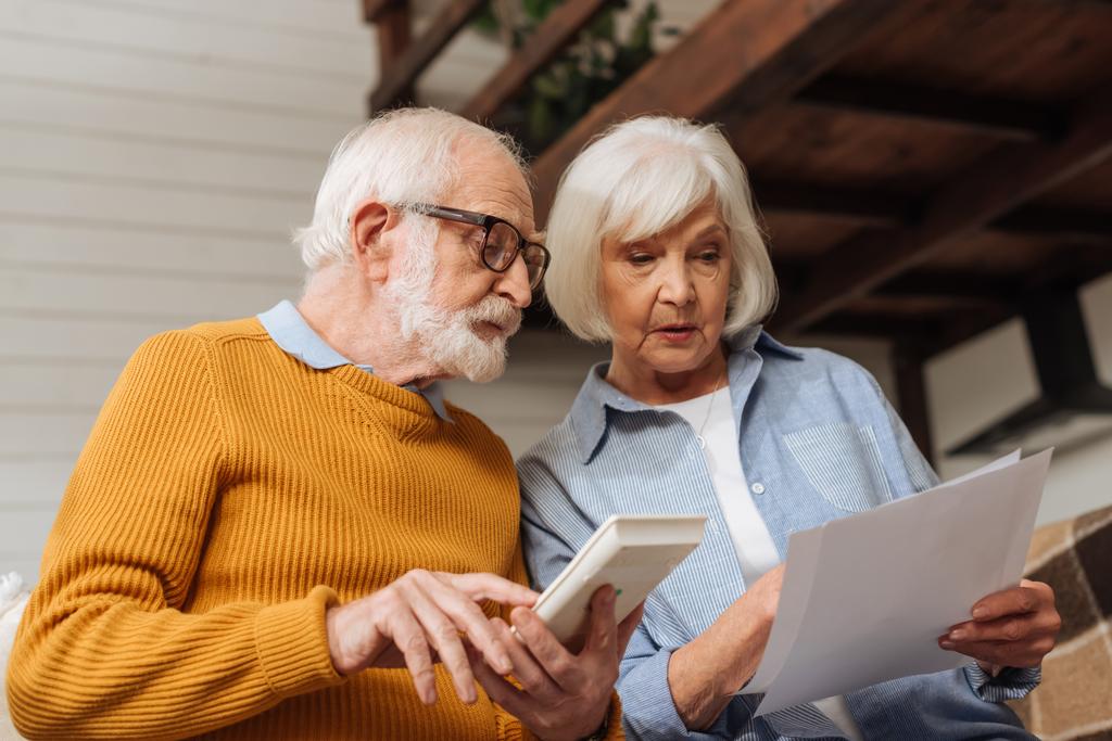 Senior Ehemann mit Taschenrechner schaut Rechnungen in der Nähe Frau auf verschwommenem Hintergrund zu Hause - Foto, Bild