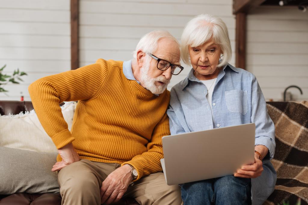 senior man met de hand op hip op zoek naar laptop in de buurt van vrouw op de bank op wazig achtergrond - Foto, afbeelding